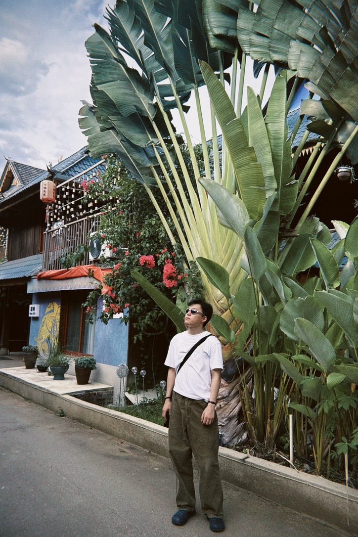 Zhou Longqing standing by tall tropical leaves and streetside architecture